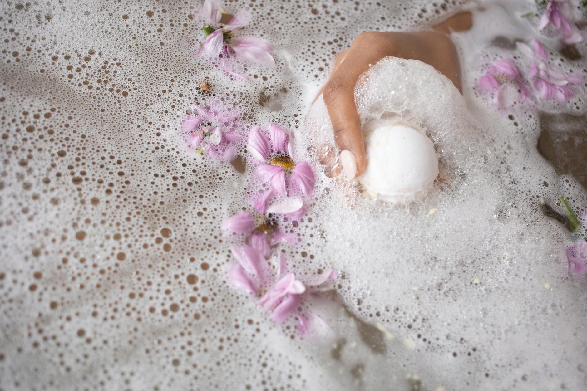 woman holding ball of bath salt in water with foam and flowers